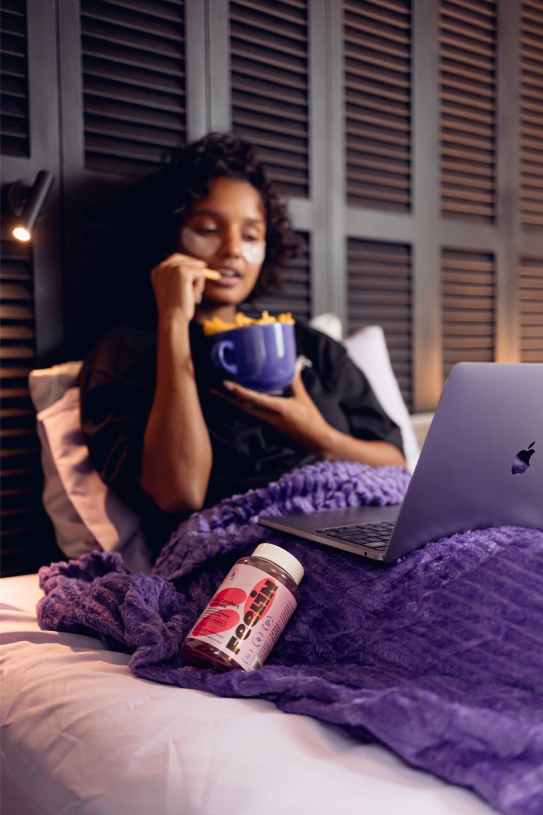 Mulher na cama com notebook, manta roxa e pote Feelin